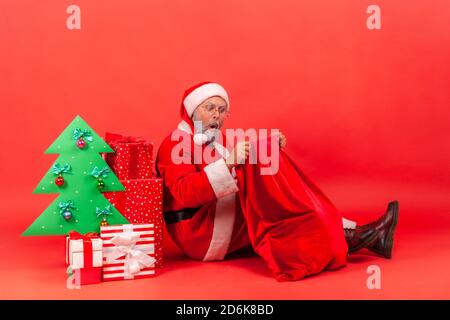 Surprised santa claus sitting on floor under decorated paper christmas tree with gift boxes and peeping into his big red bag with holidays presents. I Stock Photo