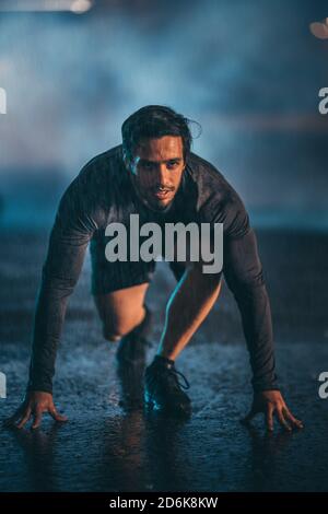 Strong Muscular Fit Young Man Ready for Sprinting on a Rainy Evening. He is Training in an Urban Environment Under a Bridge with Dark Foggy Background Stock Photo