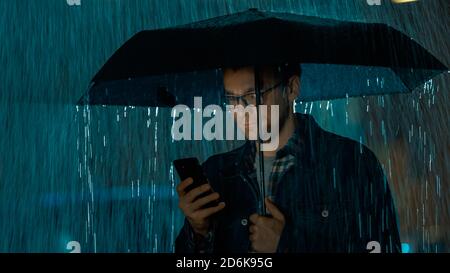 Young Caucasian Man in Glasses, Wearing a Jeans Coat and Square Shirt is Using a Smartphone Under an Umbrella. It's Dark Outside and it is Raining. Stock Photo