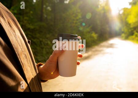 Young woman holds a disposable cup of coffee on on a sunny morning or day. Woman going for a walk with a cup of coffee on beautiful autumn day. Stock Photo