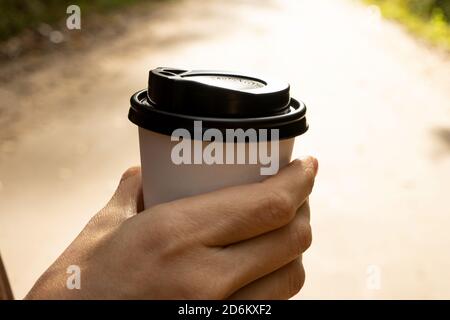 Young woman holds a disposable cup of coffee on on a sunny morning or day. Woman going for a walk with a cup of coffee on beautiful autumn day. Stock Photo