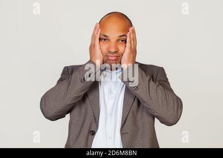 Young african american guy businessman holding his head in hands with unhappy facial expression looking sad and trying not to cry. Standing on a gray background Stock Photo