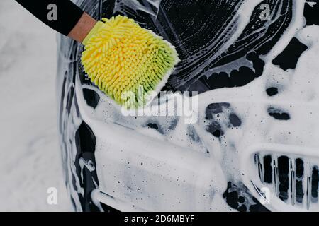 Unrecognizable man washes soapy black car with cloth at car station. Man worker cleans automobile. Vehicle and service concept Stock Photo