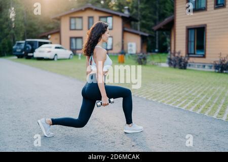 Outdoor shot of fit healthy woman dressed in active wear makes physical exercises with dumbbells in open air trains biceps poses outside with houses i Stock Photo