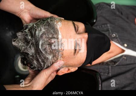 White Caucasian man with black mask of protection pandemic coronavirus is lying down while some barber's hands wash his hair with water and soap in ha Stock Photo