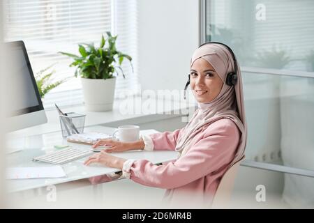 Successful young Muslim female operator in hijab and headset clicking mouse Stock Photo