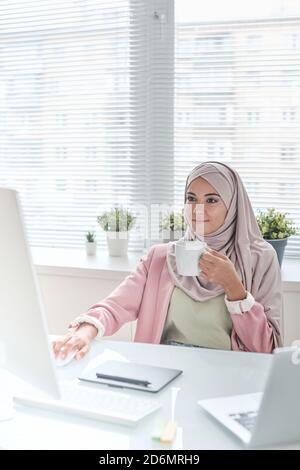 Young busy Muslim female designer or manager in hijab having tea or coffee Stock Photo