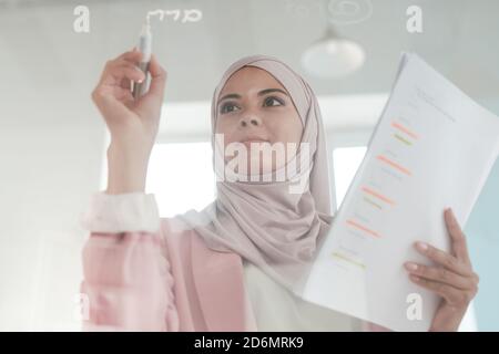 Young serious female broker in hijab holding financial papers and highlighter Stock Photo