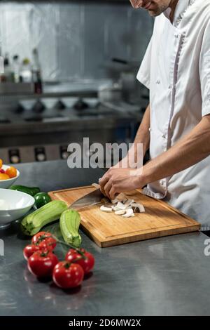 Young male chef of modern restaurant in white uniform cutting mushrooms Stock Photo