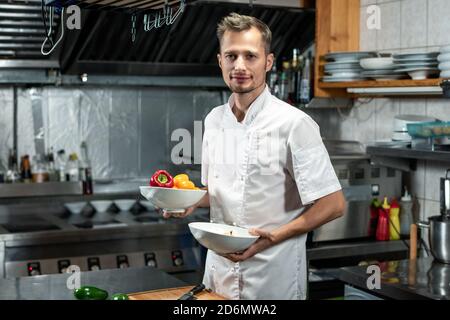 Happy young professional chef in white uniform holding bowls with fresh capsicum Stock Photo