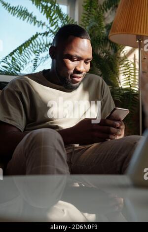 Young casual man of African ethnicity looking at smartphone screen at home Stock Photo