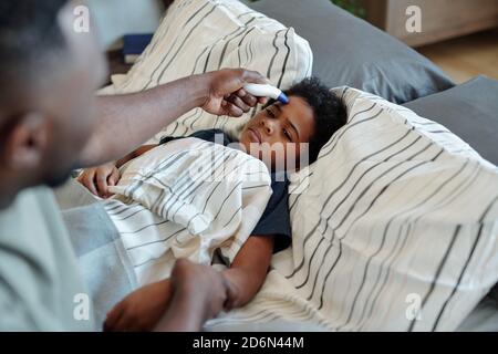 Sick little boy lying in bed and looking at his father measuring temperature Stock Photo