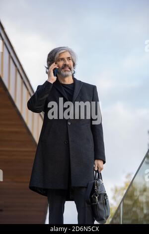 Confident grey-haired businessman in black coat making appointment by phone Stock Photo