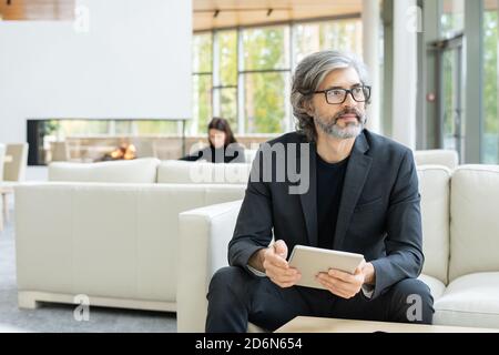 Elegant mature businessman in suit sitting on white leather couch in restaurant Stock Photo