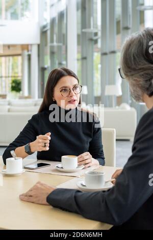Young confident businesswoman in eyeglasses sharing her ideas with colleague Stock Photo