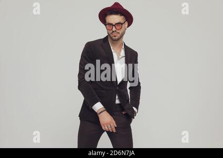 Tough fashion model holding hand in pocket, wearing hat and sunglasses while standing on gray studio background Stock Photo