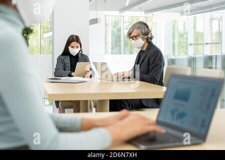 Two busy colleagues in smart casualwear and protective masks networking by table Stock Photo