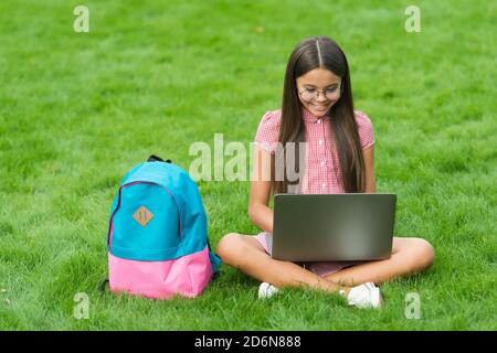 smart happy kid wear glasses using laptop for doing school homework online sitting in park on green grass with backpack, education. Stock Photo