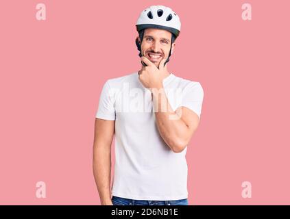 Young handsome man wearing bike helmet looking confident at the camera smiling with crossed arms and hand raised on chin. thinking positive. Stock Photo