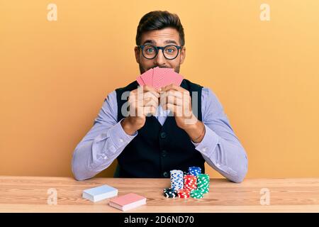 Handsome hispanic croupier man sitting on the table with poker chips and cards smiling and laughing hard out loud because funny crazy joke. Stock Photo