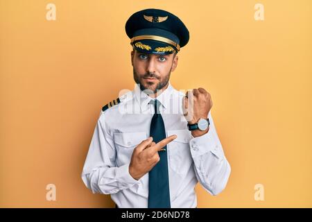 Handsome hispanic man wearing airplane pilot uniform in hurry pointing to watch time, impatience, looking at the camera with relaxed expression Stock Photo