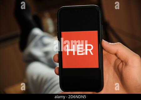 Her logo on the screen of mobile phone in mans hand. Application for dating and acquaintance. Man is laying on the sofa and looking for partner in the Stock Photo