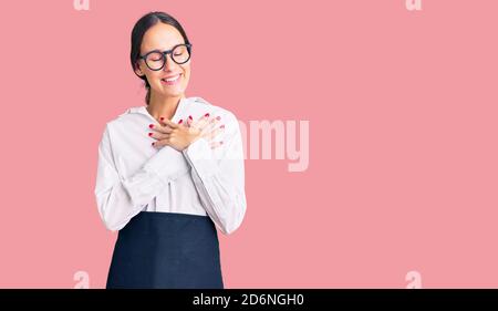 Beautiful brunette young woman wearing professional waitress apron smiling with hands on chest with closed eyes and grateful gesture on face. health c Stock Photo