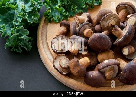 Brown boletus mushrooms on wooden cutting board and green kale leaves on table, high angle view, copy space