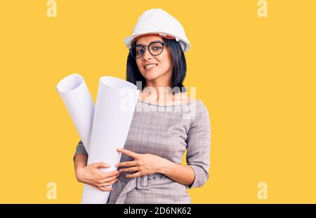 Young beautiful latin woman wearing architect hardhat and glasses holding blueprints looking positive and happy standing and smiling with a confident Stock Photo