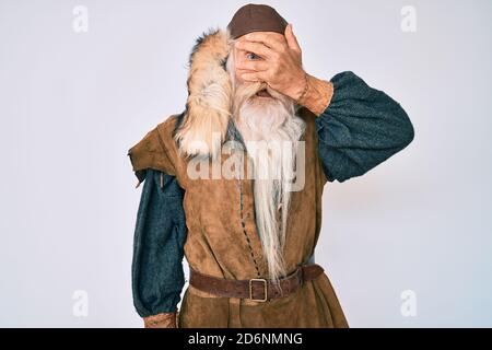 Old senior man with grey hair and long beard wearing viking traditional costume peeking in shock covering face and eyes with hand, looking through fin Stock Photo
