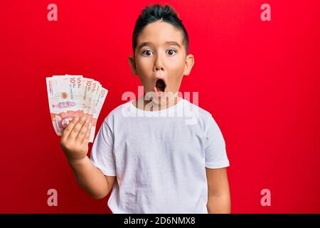 Little boy hispanic kid holding 10 colombian pesos banknotes scared and amazed with open mouth for surprise, disbelief face Stock Photo