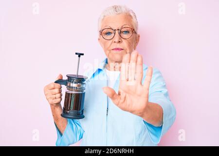 Senior beautiful woman with blue eyes and grey hair holding french coffee maker with open hand doing stop sign with serious and confident expression, Stock Photo