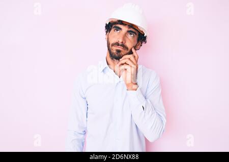 Handsome young man with curly hair and bear wearing architect hardhat thinking concentrated about doubt with finger on chin and looking up wondering Stock Photo