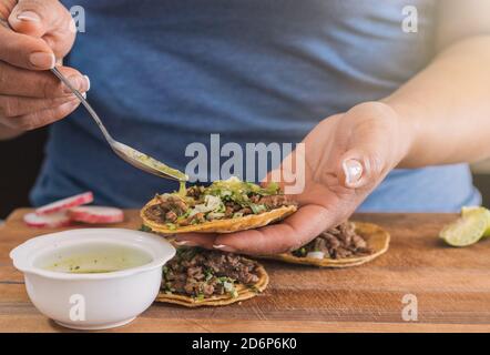 Hispanic woman adding salsa to pork tacos with yellow corn tortilla, beef, onion, lemon and green salsa. Mexican taquero preparing tacos at Mexico Stock Photo