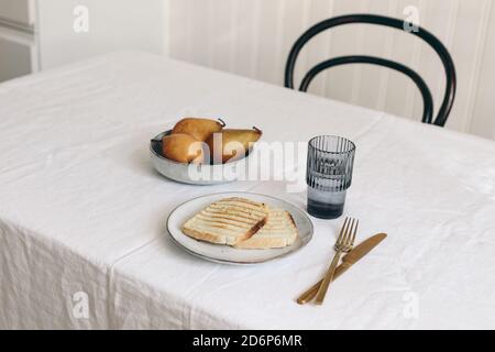 Morning breakfast with pears fruit composition. Bread toast, folded newspapers and glass of water on white linen table cloth. Minimal Scandinavian Stock Photo