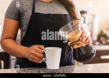 Hispanic female barista pouring coffee into a white cup in family kitchen. Black woman holding pour over coffee pot in the morning Stock Photo