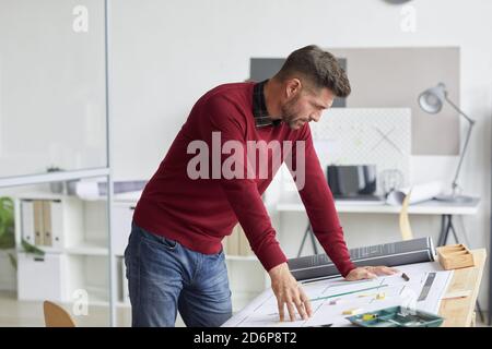 Side view portrait of bearded architect looking at blueprints while leaning on drawing table at workplace, copy space Stock Photo