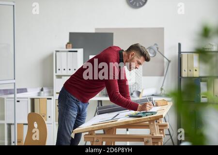 Side view portrait of bearded architect drawing blueprints while leaning on desk at workplace, copy space Stock Photo