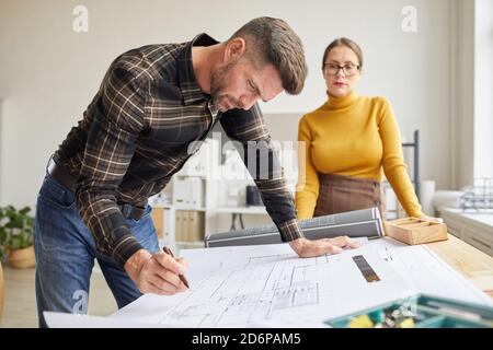 Side view portrait of mature bearded architect drawing blueprints while standing by desk in office with female colleague in background Stock Photo