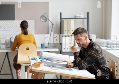 Side view portrait of mature bearded architect working on blueprints and plans while sitting at drawing desk in office, copy space Stock Photo