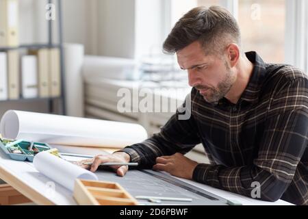 Portrait of mature bearded architect working on blueprints and plans while sitting at drawing desk in office, copy space Stock Photo