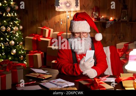 Happy Santa Claus using smartphone sitting at home table late on Christmas. Stock Photo