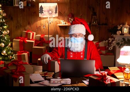 Santa wearing face mask holding Christmas gift on xmas eve using laptop. Stock Photo