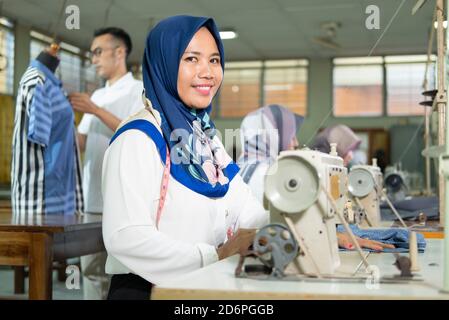 female tailor in hijab smiles look at camera with a measuring tape worn around her neck using a sewing machine while working Stock Photo