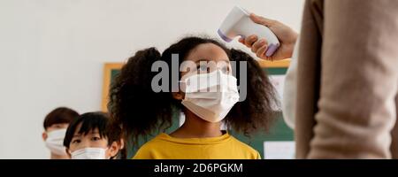 Banner group of diverse students in school building checked and scanned for temperature check. Elementary pupils are wearing face mask, line up before entering into classroom. Covid-19 school reopen Stock Photo