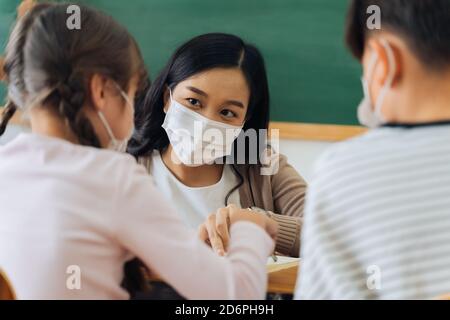 Close-up of Asian female teacher wearing a face mask in school building tutoring a primary student children. Elementary pupils writing and learning in classroom. Covid-19 school reopen concept Stock Photo