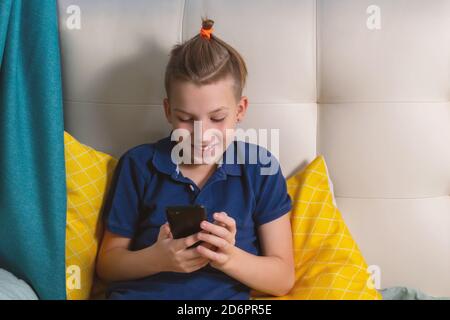 Caucasian boy sitting on bed , smiling, holding smartphone in hands and talking using video call with friends. Friendship in new reality during lockdo Stock Photo