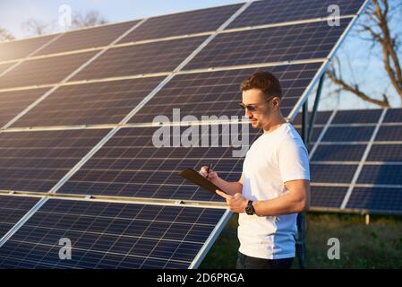 Portrait of a young successful man standing by a solar pv plant looking in his folder pondering on some business plan and making notes, side view, alternative energy concept Stock Photo