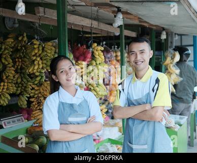 Smiling female and male shopkeeper in apron standing with crossed hands in the fruit market Stock Photo