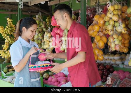 A smiling female shopkeeper in apron give service for male customers who want to buy fruit parcels in the fruit market Stock Photo